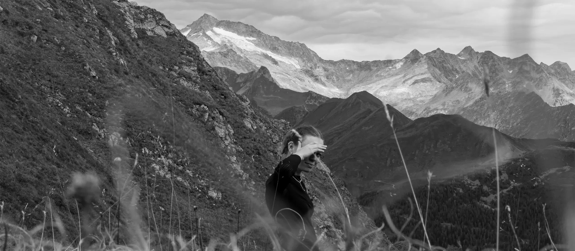 Karen steht halb durch Grashalme verdeckt in einer Berglandschaft. Im Hintergrund sind weitere Berge mit Schnee zu sehen.