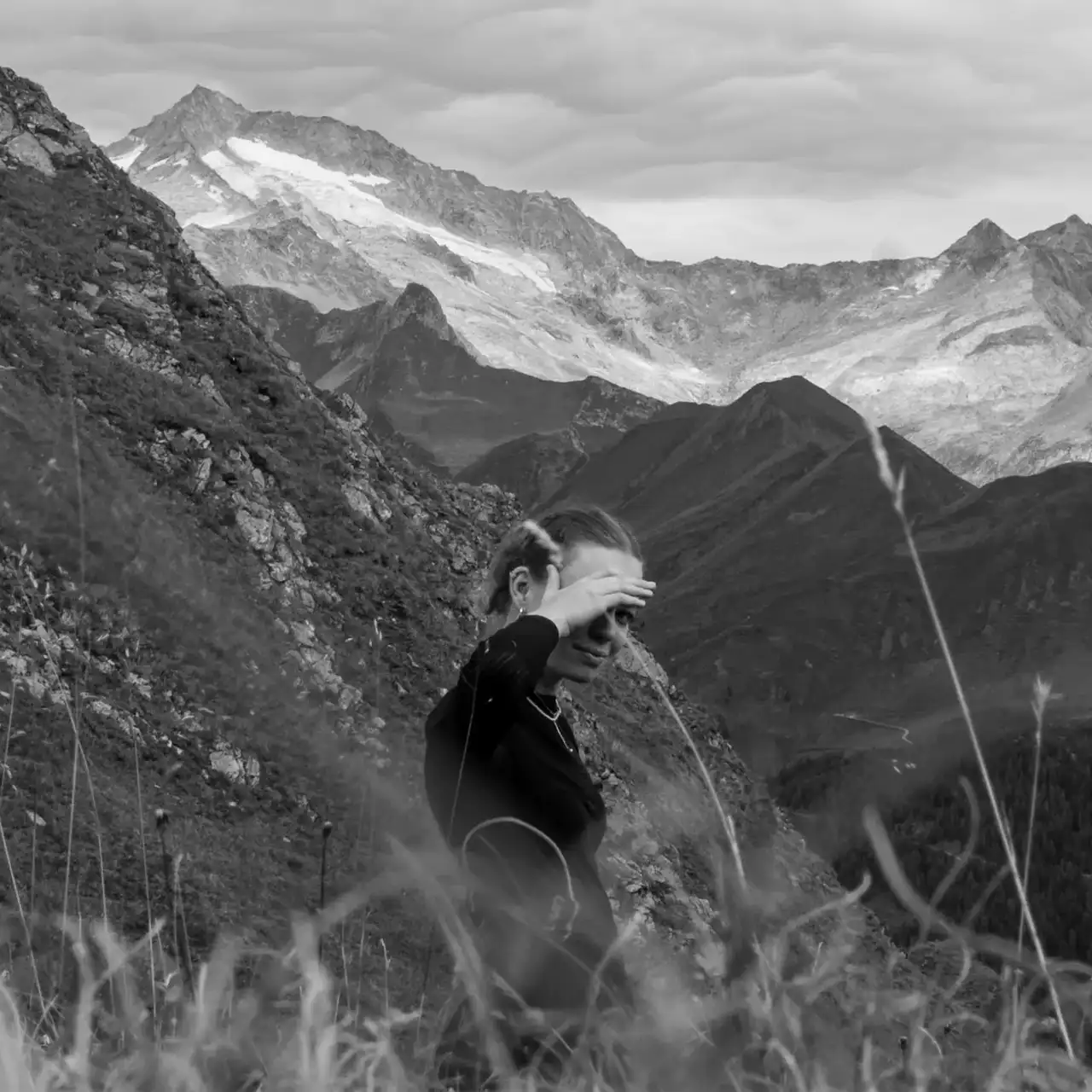 Karen steht halb durch Grashalme verdeckt in einer Berglandschaft. Im Hintergrund sind weitere Berge mit Schnee zu sehen.