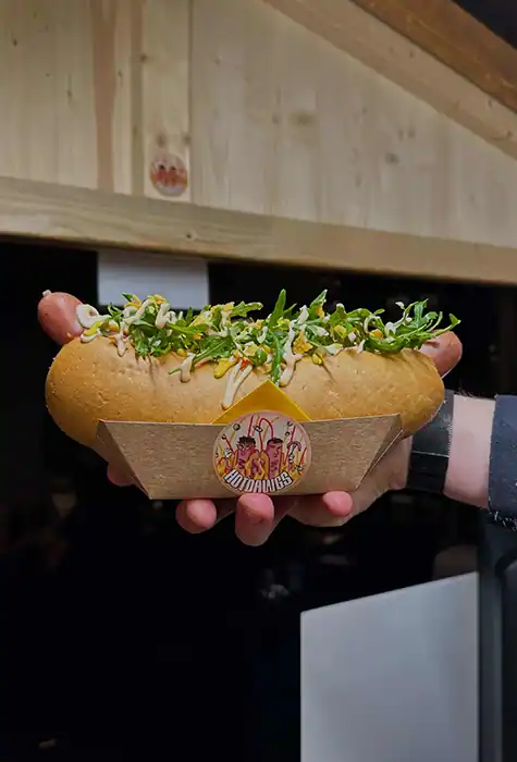 Eine Hand hält einen Hotdog. Auf der Verpackung ist der Hotdawg-Sticker zu sehen.