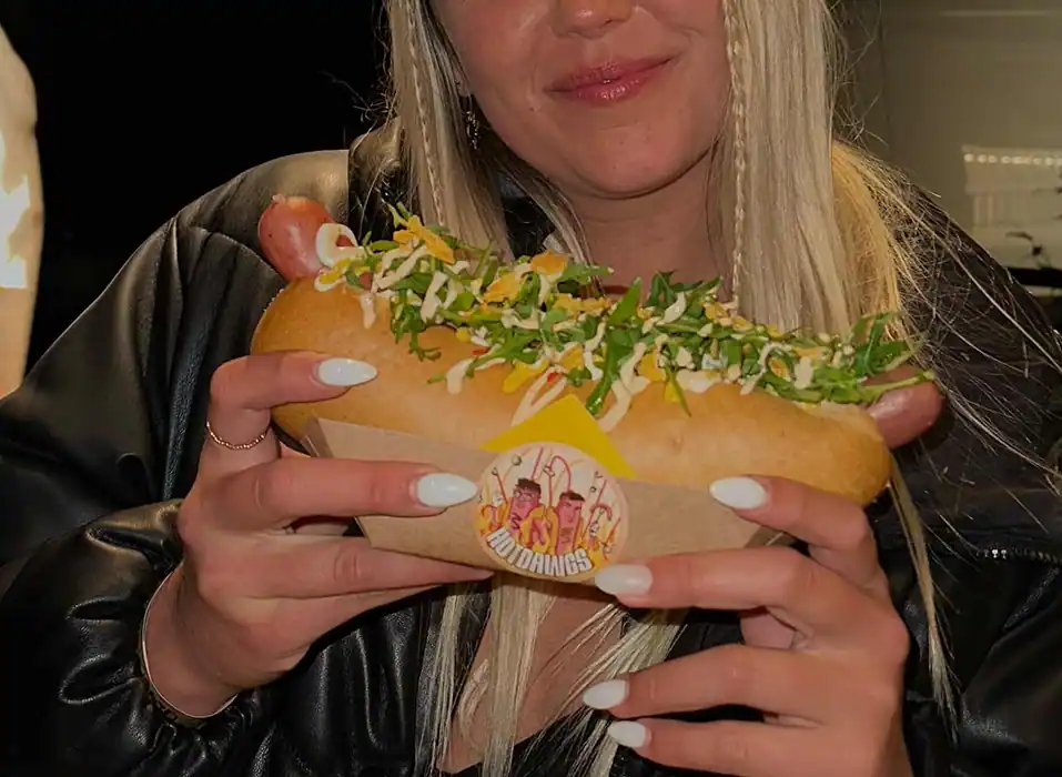 Eine junge Frau hält lächelnd einen Hotdog in der Hand. Auf der Verpackung ist der Hotdawg-Sticker zu sehen.